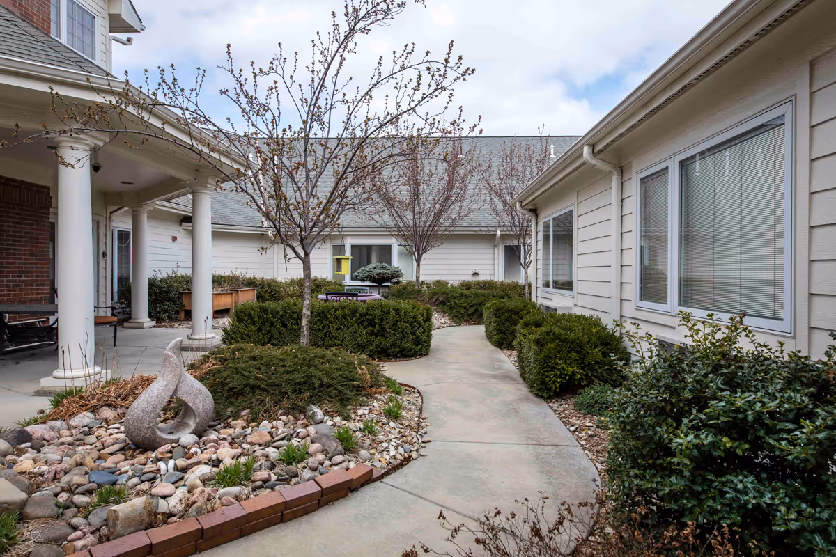 Outdoor courtyard area at Pioneer Ridge Independent Living of Lawrence featuring a curved concrete pathway surrounded by bushes, small trees with budding leaves, and a rock garden with a modern stone sculpture. The courtyard is bordered by light-colored buildings with windows and a covered porch with white columns.