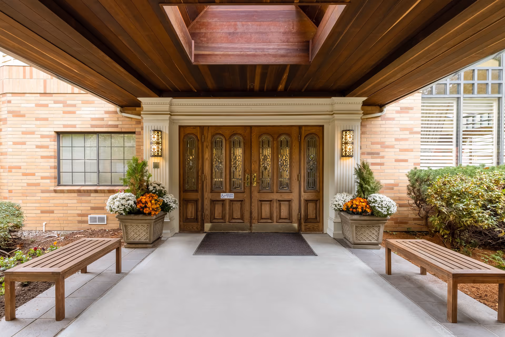 Entrance to a senior living facility with a wooden double door featuring decorative glass panels, flanked by two wall-mounted lantern-style lights. There are two wooden benches on either side of the entrance and large planters with white and orange flowers and greenery. The building exterior is made of light-colored brick, and the entrance area has a wooden ceiling with a skylight.
