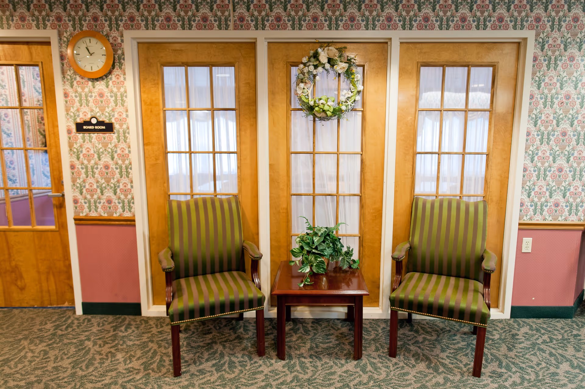 A waiting area with two green and brown striped armchairs separated by a small wooden table with a green potted plant. Behind the chairs are three wooden-framed glass panels with white curtains, and a floral wreath hangs on the middle panel. To the left, there is a wooden door with a sign that reads 'Board Room' and a round wall clock above it. The walls have floral wallpaper with a pink lower section and green carpeted floor.