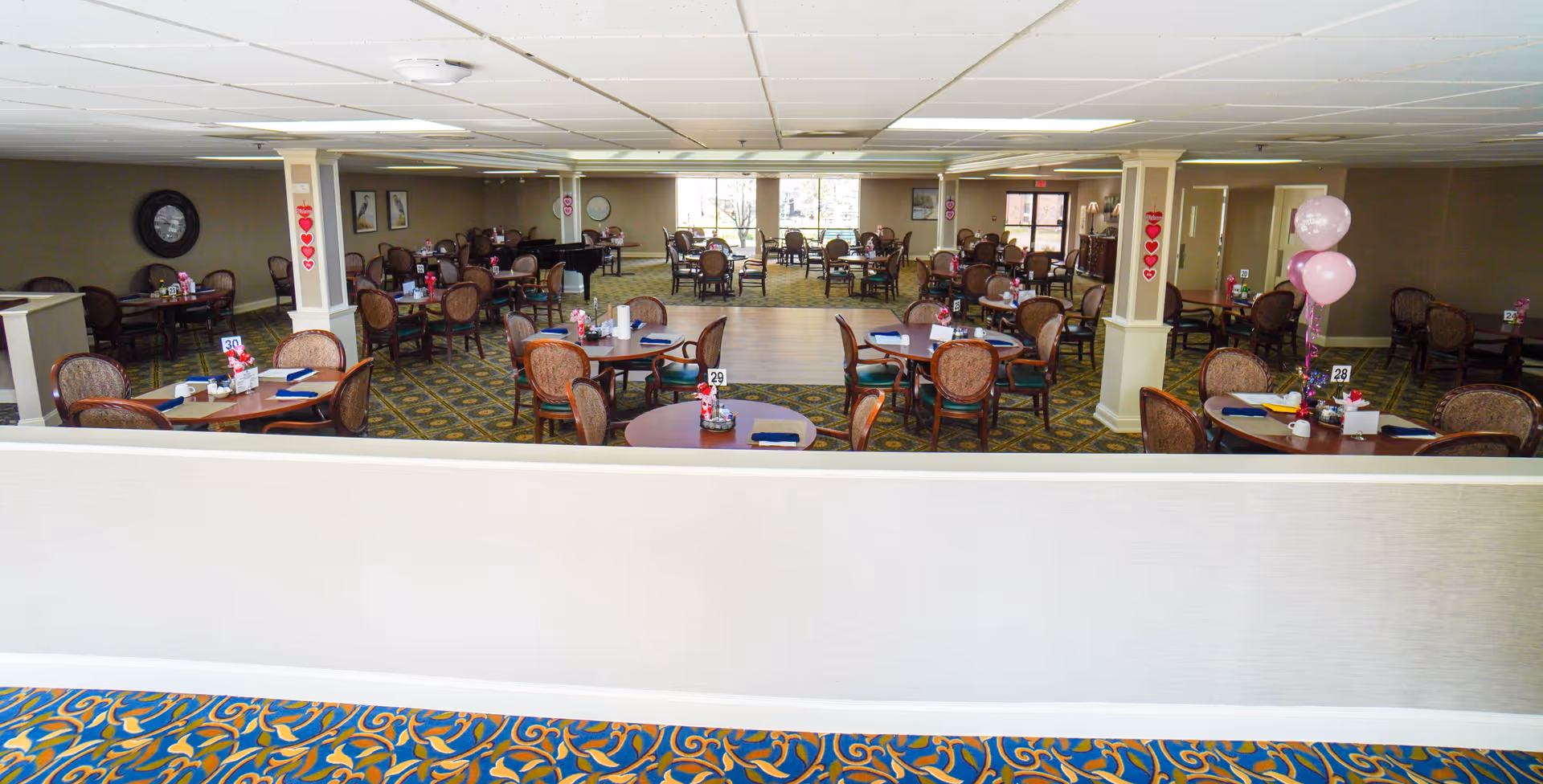 Spacious dining room with many round tables and chairs set up and decorated with table numbers and balloons.