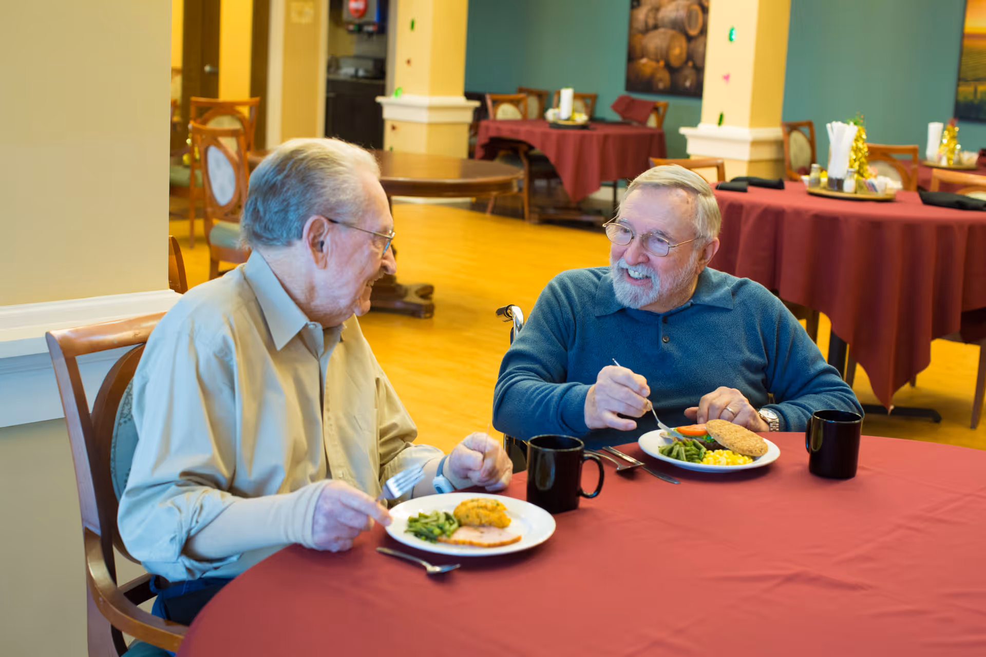 Two elderly men sit at a table in a dining room, smiling and eating meals.