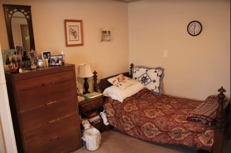 A small bedroom with a single wooden bed covered with a patterned red bedspread and multiple pillows. Next to the bed is a wooden nightstand with a lamp, books, and other items. To the left is a tall wooden dresser with framed photos and decorative items on top. A wall clock is mounted on the plain beige wall above the bed.