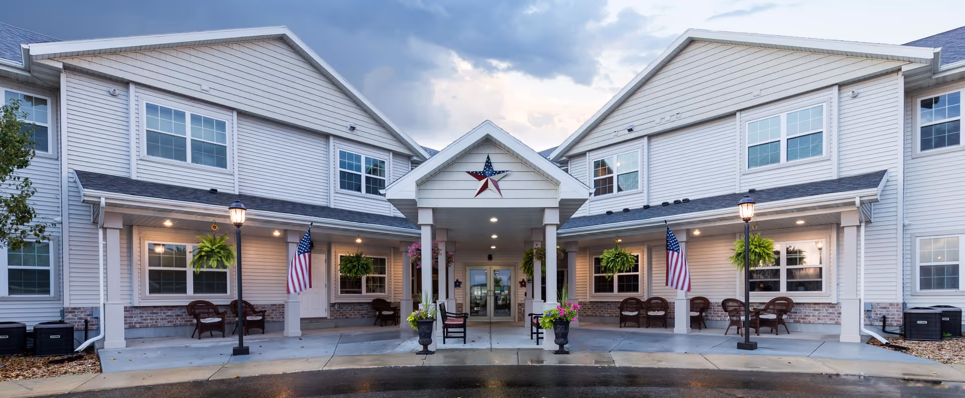 Front exterior view of The Meadows Of Fall River, L.L.C. facility showing a two-story building with white siding, multiple windows, a covered entrance with columns, American flags, hanging plants, outdoor seating, and lampposts under a cloudy sky.