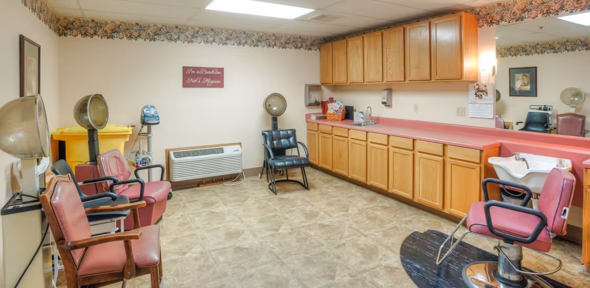 Interior view of a vintage-style hair salon with pink salon chairs, hair dryers, a sink for washing hair, wooden cabinets with a pink countertop, and a wall-mounted air conditioning unit. The room has a floral wallpaper border near the ceiling and a sign on the wall that reads 'I'm a Beautician Not a Magician.'