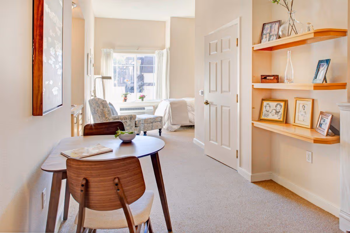 A bright and cozy room in a senior living facility with a small wooden table and two chairs in the foreground. The table has a bowl of green apples and a magazine. In the background, there is a comfortable armchair with floral upholstery near a window with white curtains. A bed with white bedding is partially visible. On the right wall, there are three wooden shelves displaying framed photos and decorative items.
