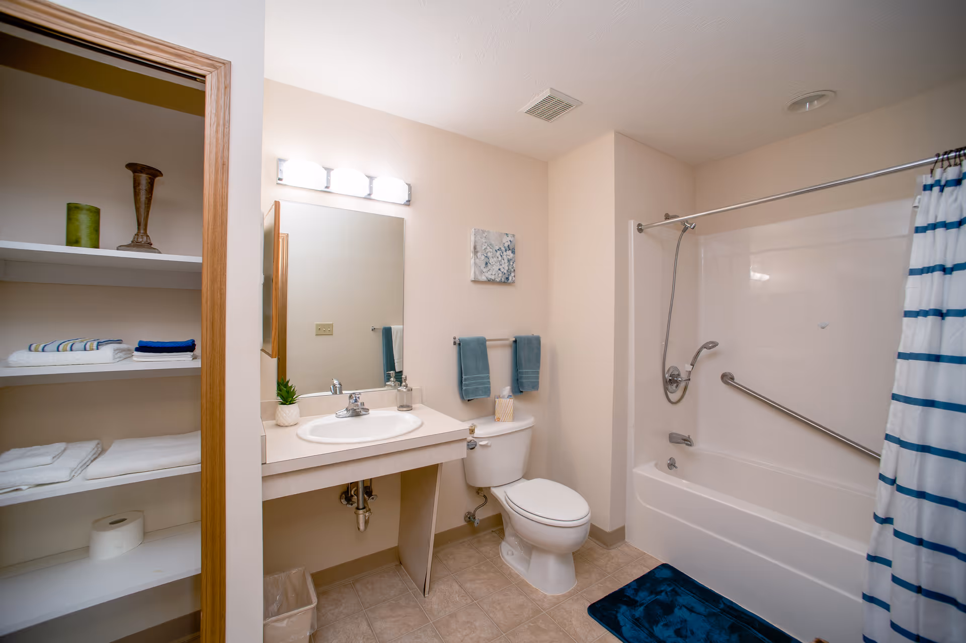 Accessible senior bathroom featuring a sink and mirror, toilet, bathtub with grab bar and striped shower curtain, and open linen shelves.