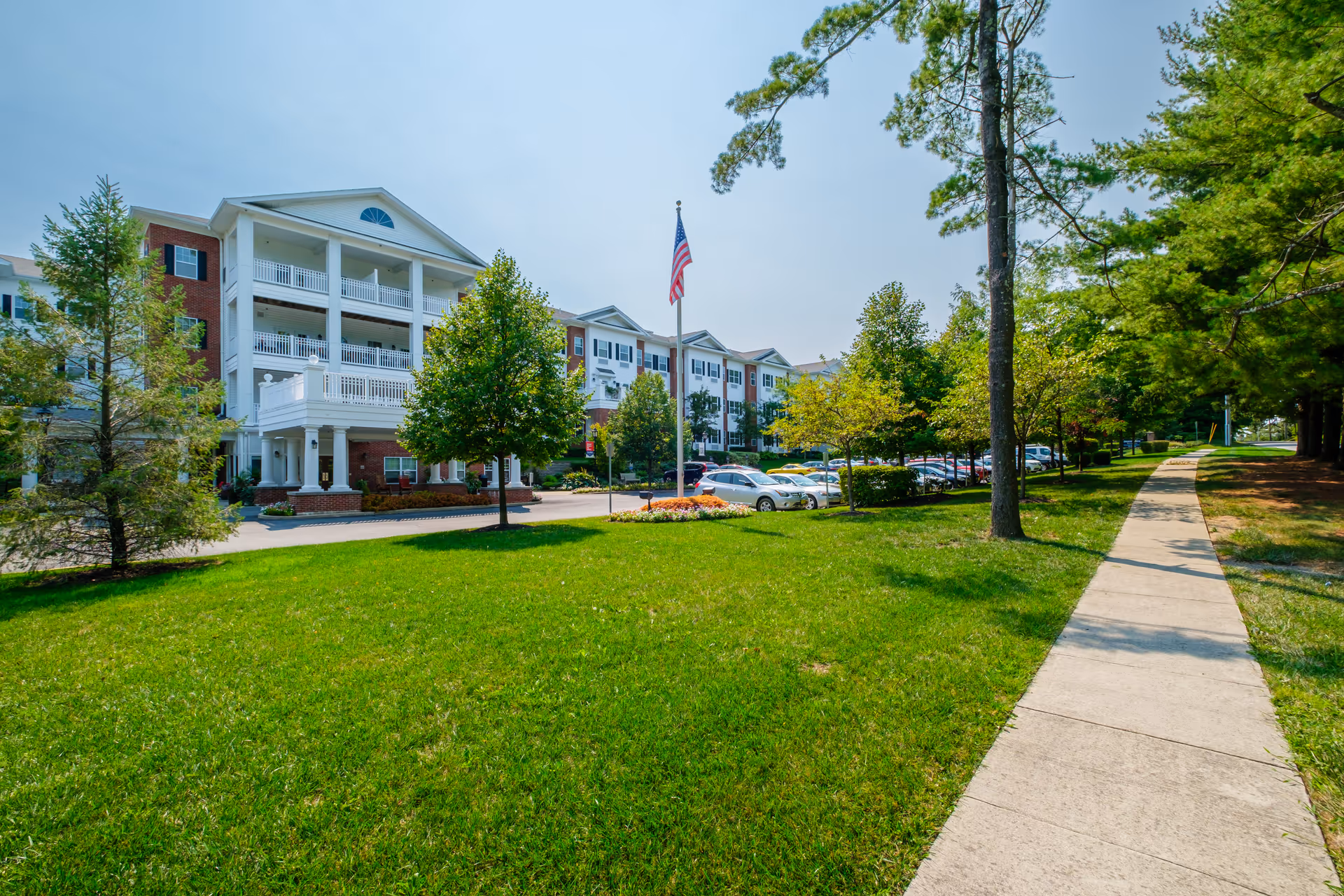 Exterior view of a senior living facility with a large green lawn, trees, a sidewalk, and a parking lot with cars. The building is multi-story with white balconies and brick accents. An American flag is prominently displayed on a flagpole in front of the building.