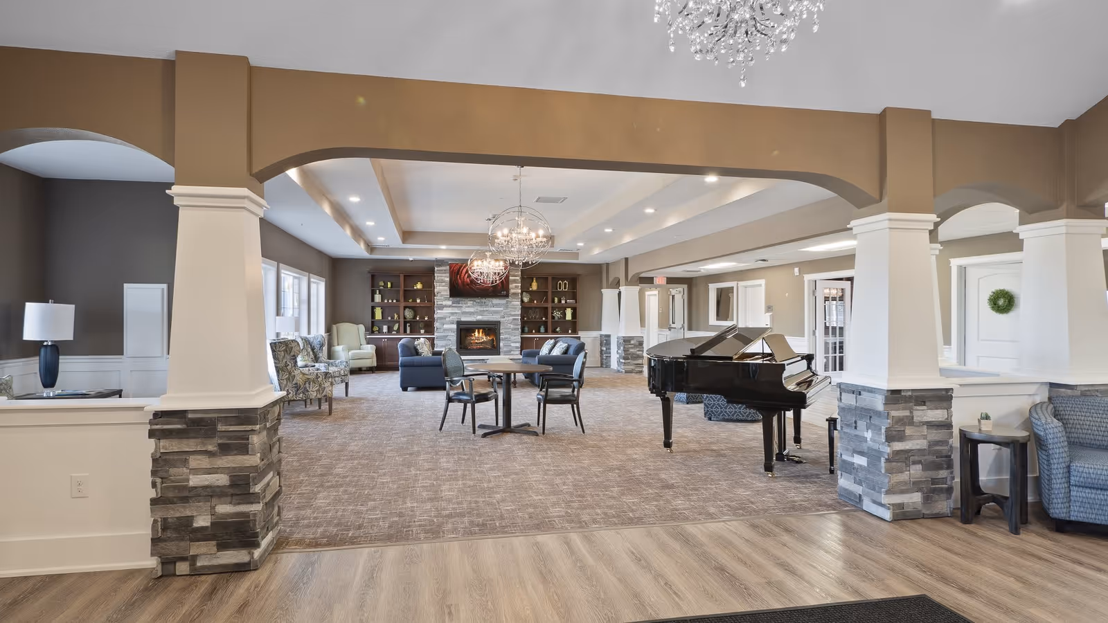 Spacious and elegant living room area with a grand piano on the right side, a round table with four chairs in the center, and a cozy seating area with armchairs near a stone fireplace and built-in shelves in the background. The room features beige walls, stone pillars, wood flooring in the foreground, carpeted area in the seating space, and modern chandeliers hanging from the ceiling.