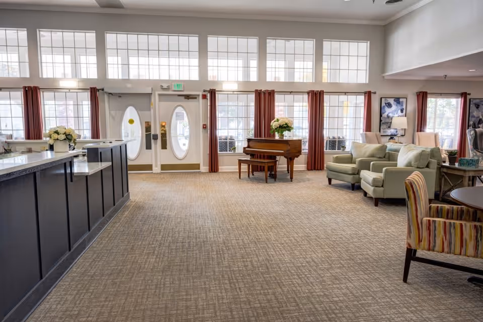 Spacious and well-lit senior living facility common area with large windows, a reception desk on the left, a grand piano with a flower arrangement in the center, and comfortable seating including armchairs and a striped chair on the right.
