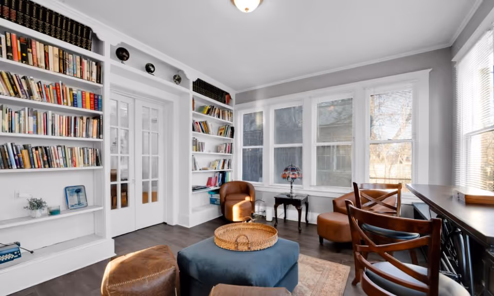 Sunlit living room with built-in bookshelves, multiple chairs, a small table and large windows.