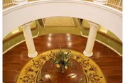 View from above of a circular wooden table with a floral centerpiece on a patterned round rug, surrounded by white columns and a curved staircase with carpeted steps and a wooden handrail.