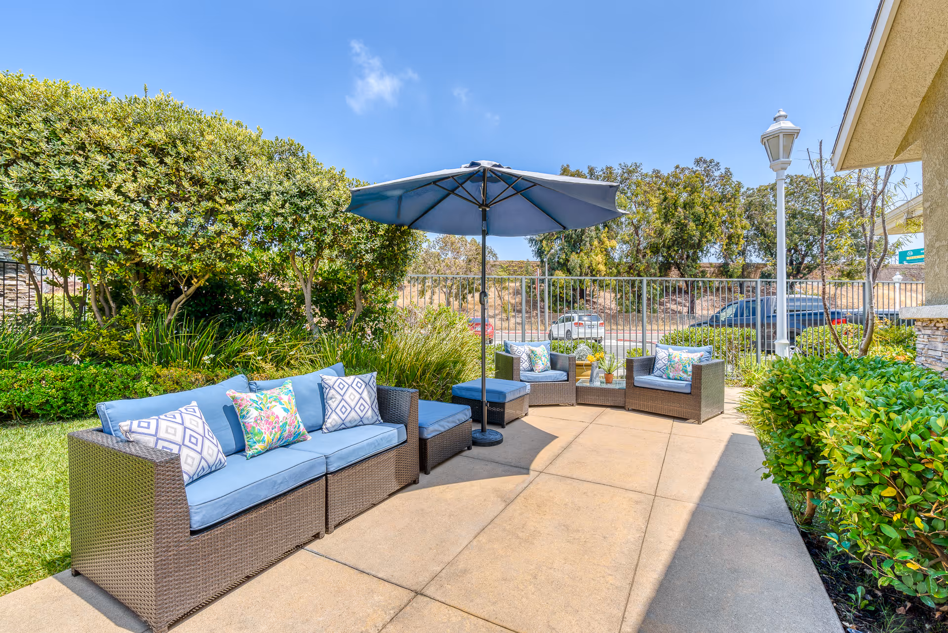 Outdoor patio area at Newport Mesa Senior Living with wicker furniture including a sofa, chairs, and ottomans with blue cushions and decorative pillows. A large blue umbrella provides shade. The patio is surrounded by green bushes and trees, with a metal fence and parked cars visible in the background under a clear blue sky.