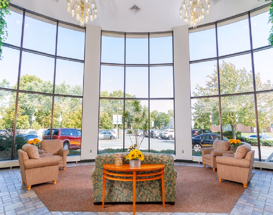 A bright and spacious seating area with large floor-to-ceiling windows showing a parking lot and trees outside. The room features two beige armchairs on each side, a floral patterned sofa in the center with a wooden table behind it holding a basket of yellow flowers and a small decorative sign. The floor is tiled around the seating area, which is covered by a large carpet. Two chandeliers hang from the ceiling.