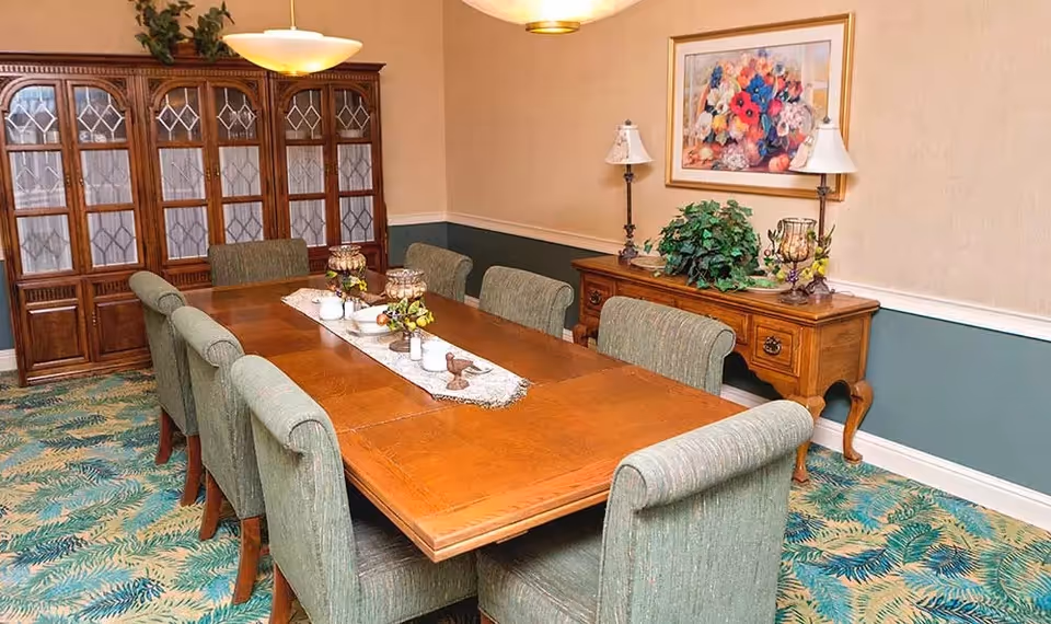 A dining room with a long wooden table surrounded by eight upholstered chairs. The table is decorated with a lace runner and small floral centerpieces. Against the wall is a wooden sideboard with two lamps, a plant, and a framed floral painting above it. A large wooden cabinet with glass doors is visible in the background. The carpet has a green and blue leaf pattern.