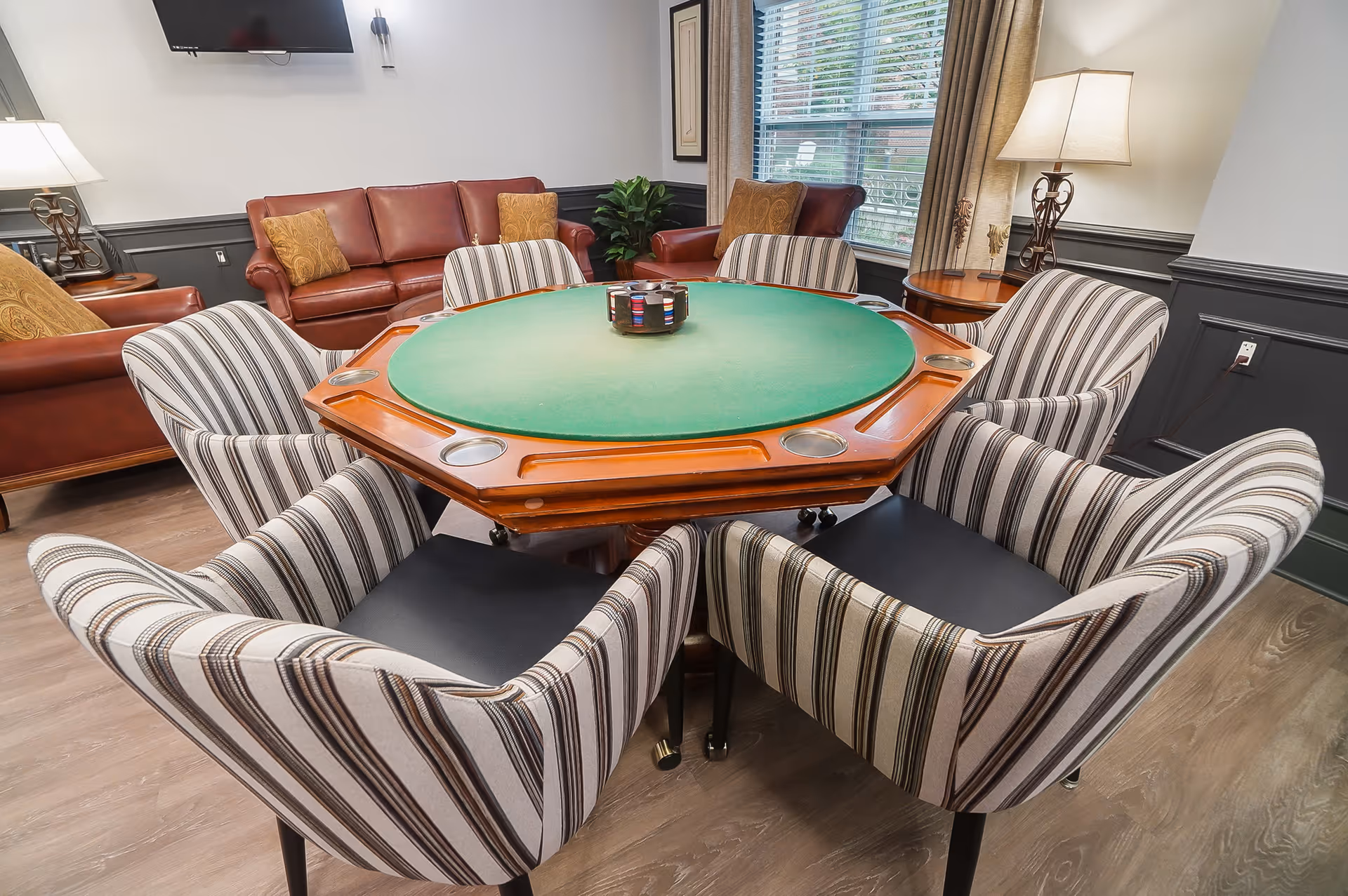 A cozy common area with a green felt octagonal poker table surrounded by six striped upholstered chairs. In the background, there are brown leather sofas with decorative pillows, a side table with a lamp, a large window with blinds, and a wall-mounted TV.