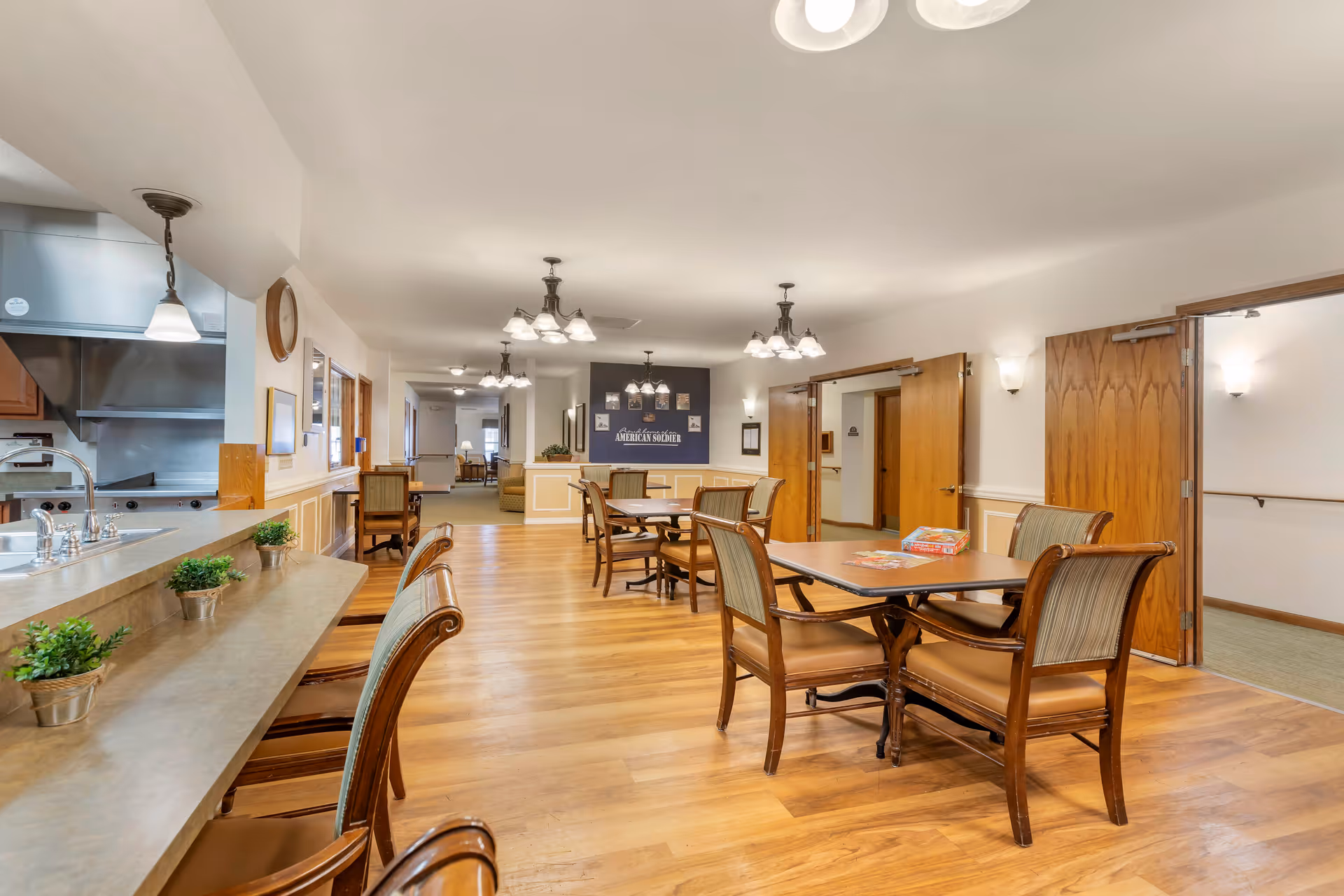 A spacious dining area with wooden floors and multiple tables and chairs arranged neatly. The room features warm lighting with chandeliers and wall sconces. On the left side, there is a counter with a sink and small potted plants. The walls are painted white with wooden trim, and there are open double doors leading to a hallway. A sign on the far wall reads 'Brookdale home of the AMERICAN SOLDIER'.