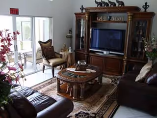 A cozy living room with a dark leather sofa and armchair, a wooden coffee table on a patterned rug, a large wooden entertainment center with a flat-screen TV, and glass doors leading to an outdoor patio area. The room is decorated with flowers and small decorative items.