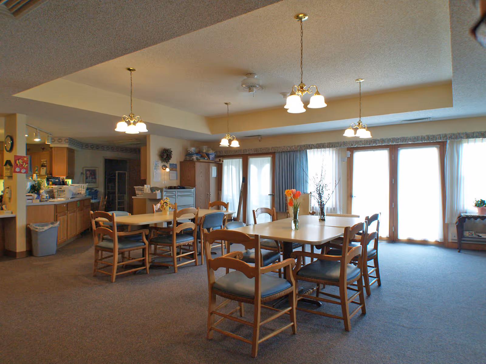 A dining room in a senior living facility with multiple wooden tables and chairs arranged neatly. The room is well-lit with hanging light fixtures and natural light coming through large windows and glass doors covered with sheer curtains. There are flower vases on the tables and a kitchen area visible in the background.