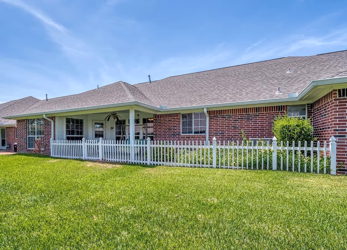 Exterior view of a single-story brick building with a gray shingled roof, white picket fence, green lawn, and a covered porch area with ceiling fans under a clear blue sky.