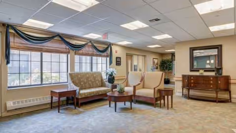 A spacious senior living lounge with sofa, chairs, tables, and large windows.