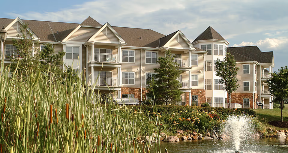 Exterior view of a multi-story senior living facility named Immanuel: Lakeside, featuring beige siding with brick accents, balconies, and a landscaped garden with tall grasses, trees, flowers, and a pond with a water fountain in the foreground.