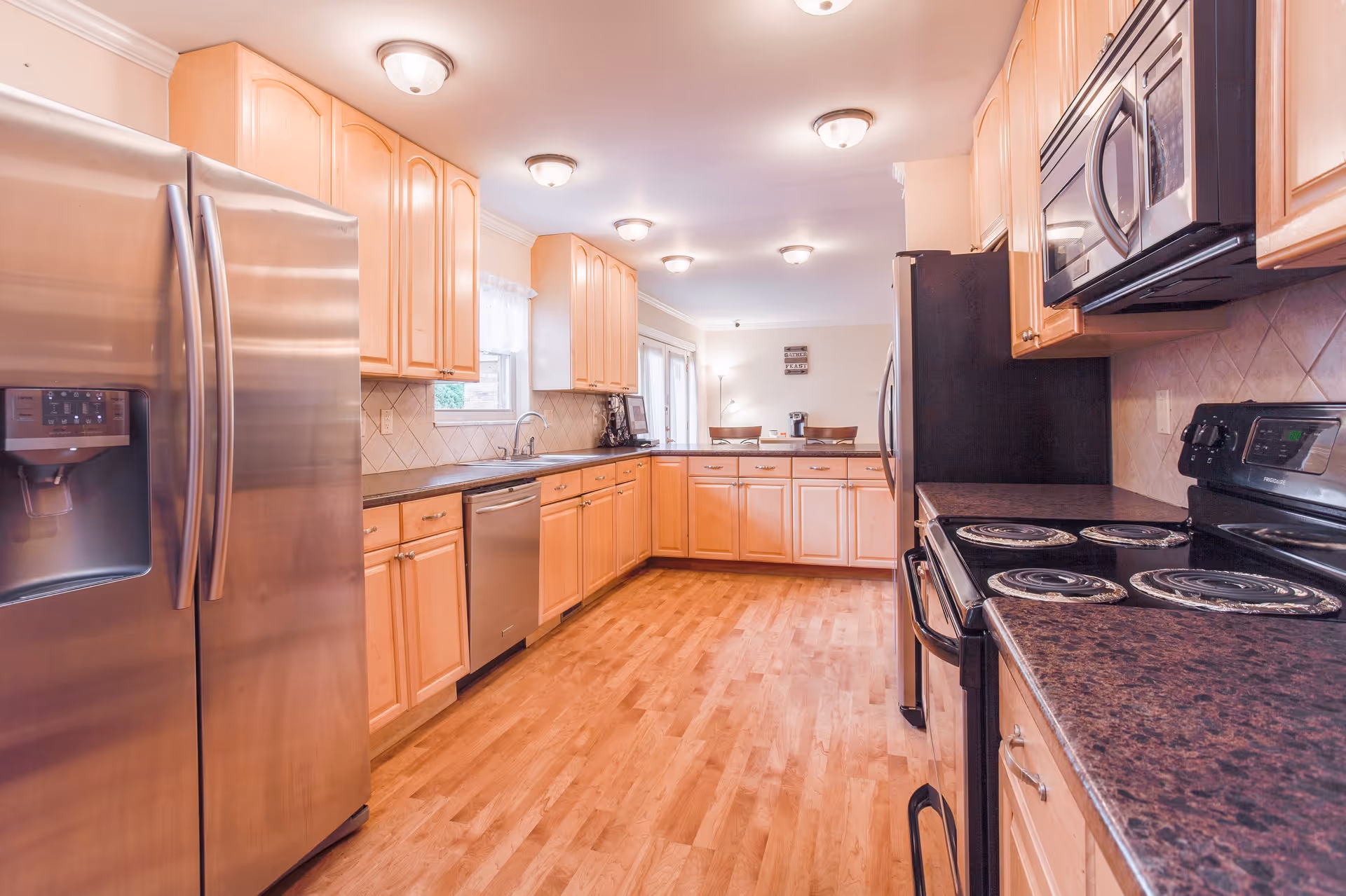 A spacious kitchen with wooden cabinets, stainless steel refrigerator and dishwasher, black electric stove, microwave, and brown countertops. The kitchen has wooden flooring and multiple ceiling lights. In the background, there is a small dining area with two chairs and a coffee maker on the counter.
