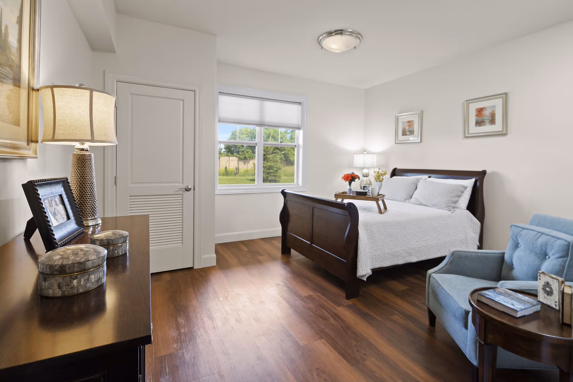 A bright and clean bedroom with a wooden bed frame, white bedding, two framed pictures on the wall, a blue armchair next to a round wooden side table with books, a wooden dresser with a lamp and photo frame, and a window showing greenery outside.