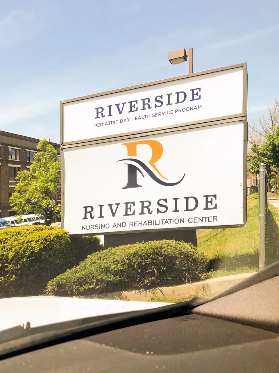 Outdoor sign for Riverside Nursing and Rehabilitation Center and Riverside Pediatric Day Health Service Program, with bushes and a building in the background.