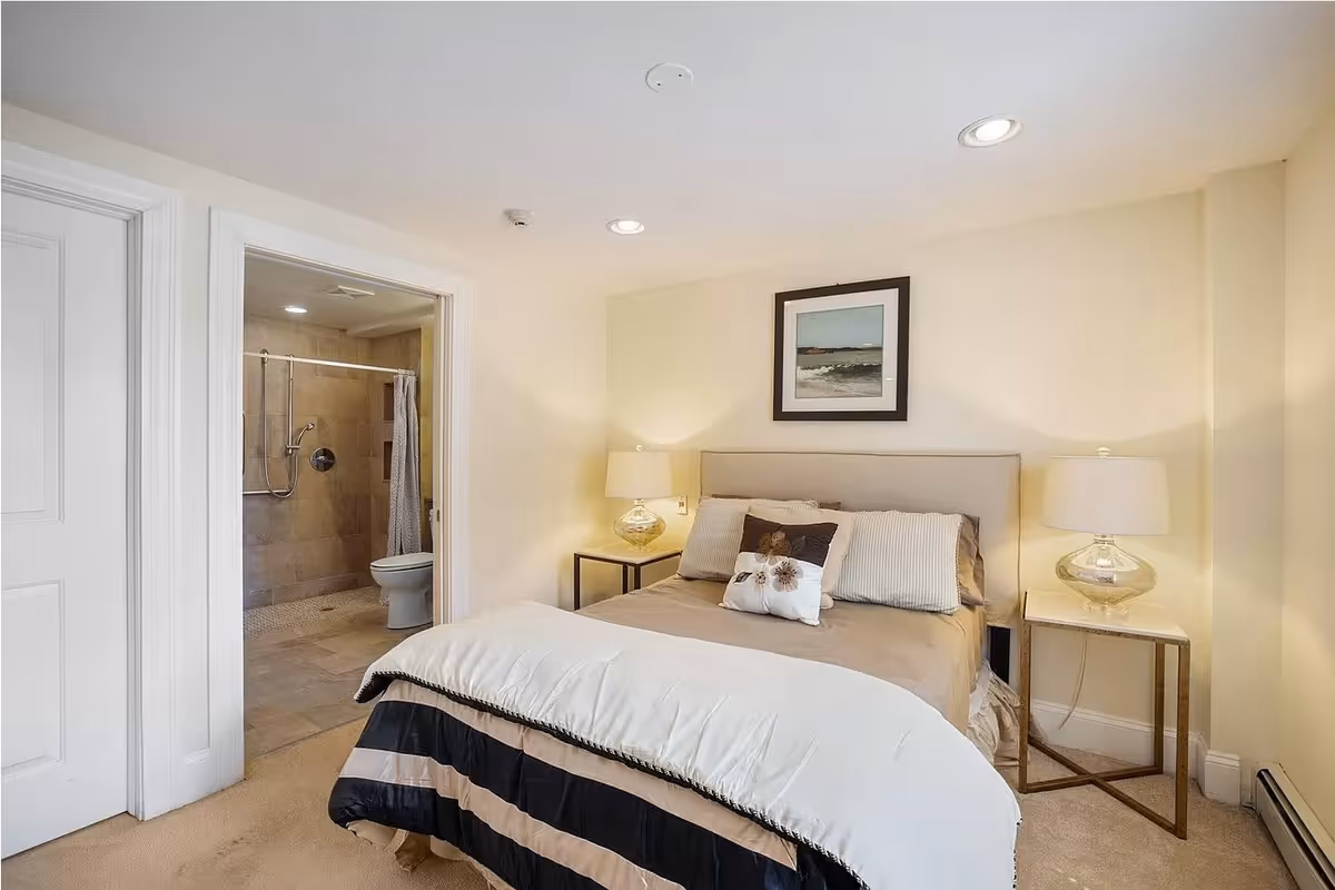 A cozy bedroom with a neatly made bed featuring multiple pillows and a beige headboard. Two bedside tables with matching lamps flank the bed. A framed picture hangs on the wall above the headboard. To the left, an open door reveals a bathroom with a walk-in shower and a toilet.