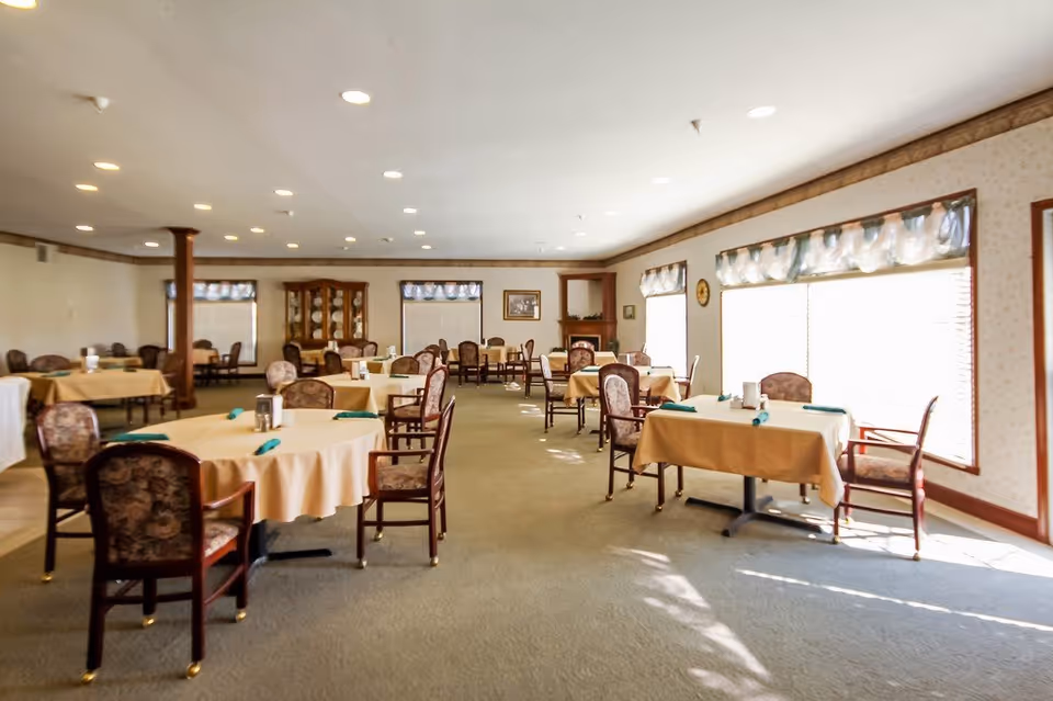 Bright dining room with round and rectangular tables covered in tablecloths and chairs arranged around them in a senior living facility.