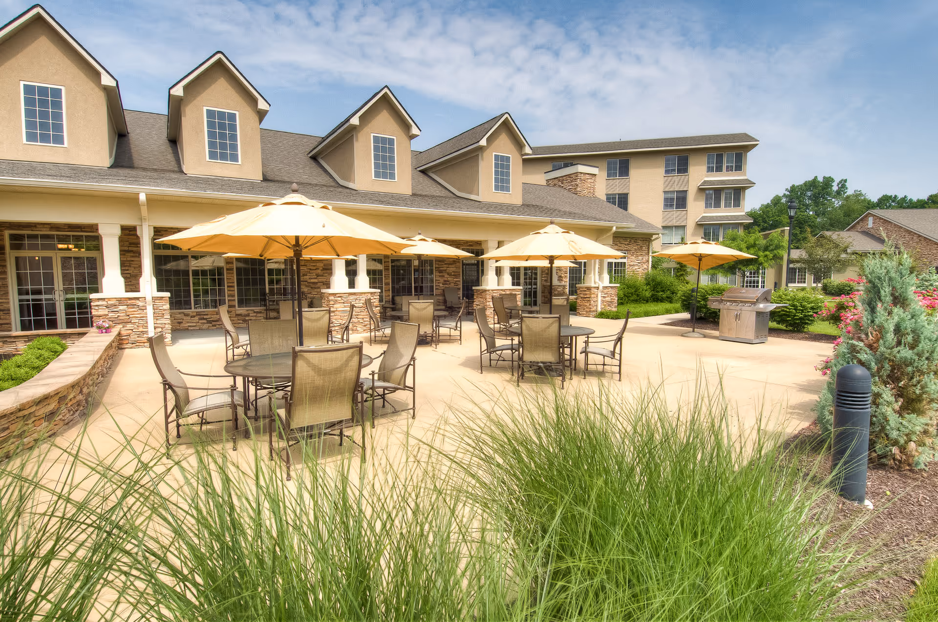 Outdoor patio area at Westside Garden Plaza featuring multiple round tables with beige umbrellas and chairs, surrounded by greenery and landscaping. The building with stone and beige exterior walls is visible in the background under a partly cloudy sky.