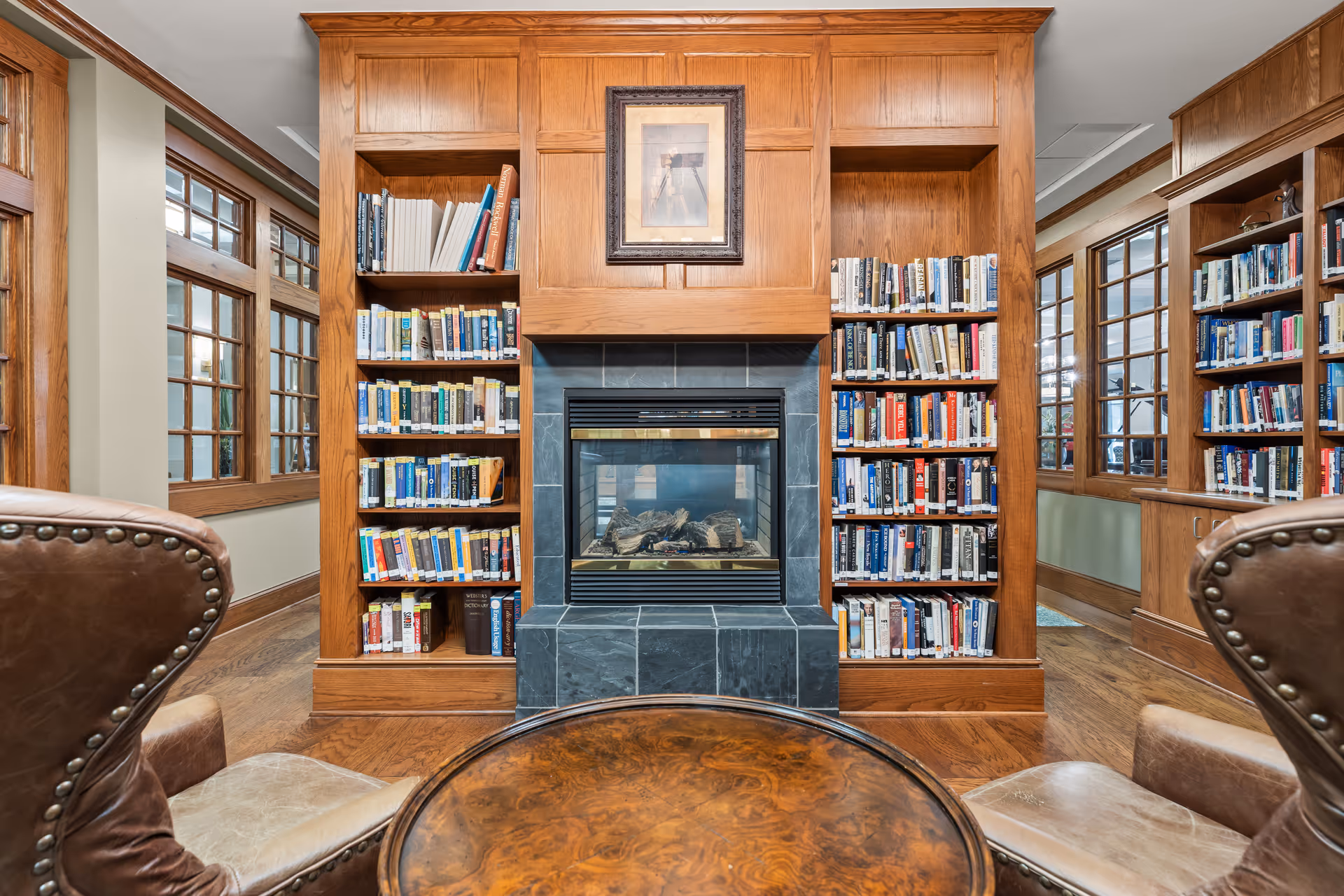 Wood-paneled library with built-in bookshelves flanking a central fireplace, leather chairs, and a round wooden table in the foreground.