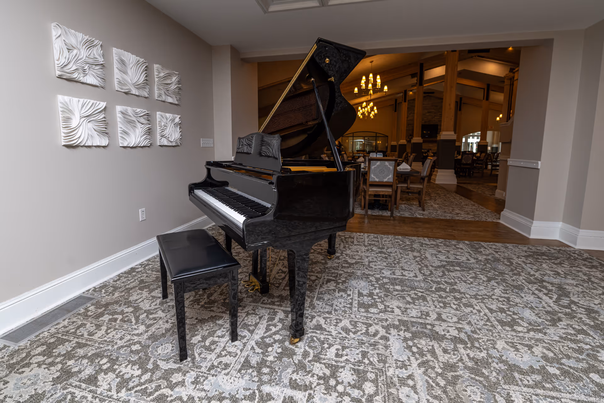 A black grand piano with a matching bench sits on a patterned carpet in a spacious room. The walls are light-colored and decorated with six white, textured square art pieces arranged in two rows. In the background, there is a dining area with tables and chairs, illuminated by chandeliers hanging from a high ceiling.