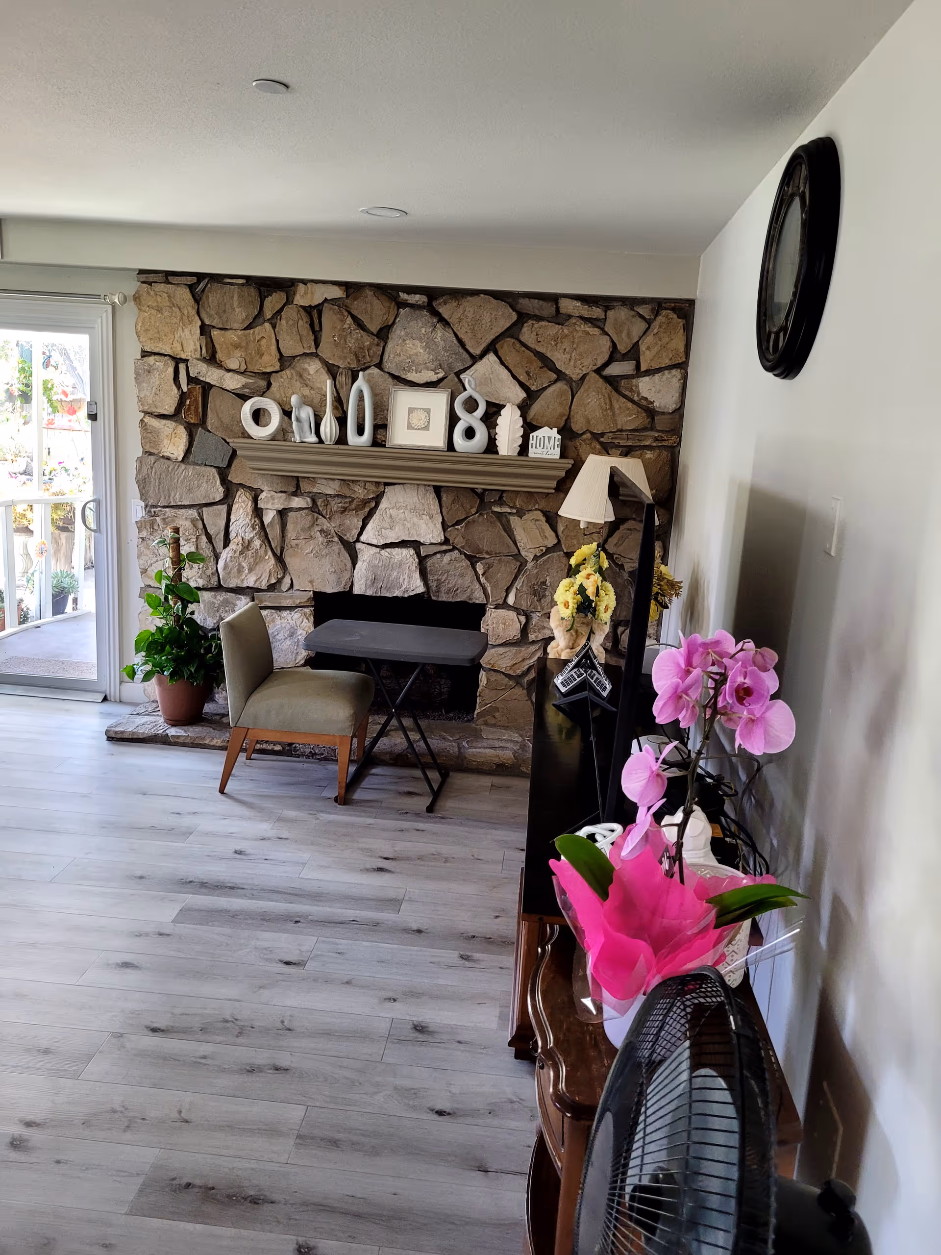 A cozy living room with a stone fireplace as the focal point. The fireplace mantel is decorated with various white decorative items and a small framed picture. In front of the fireplace is a small table with a chair. To the right, there is a wooden cabinet with a lamp, yellow flowers, and a pink orchid plant. A large wall clock is mounted on the white wall above the cabinet. The floor is light-colored wood, and a glass sliding door on the left leads to an outdoor area with plants.