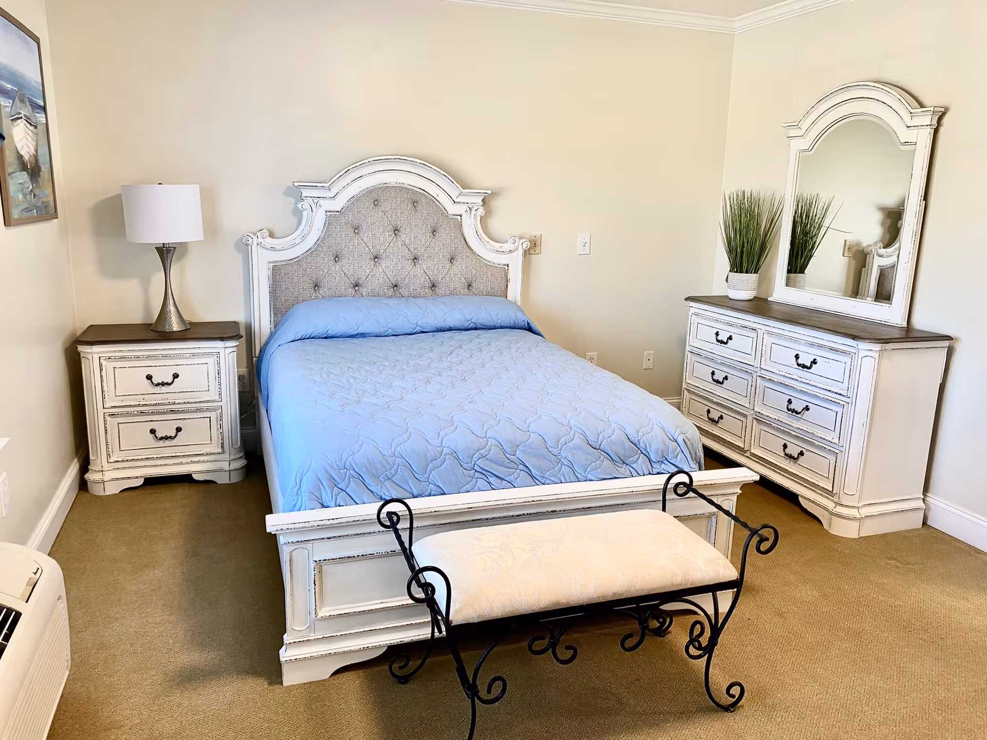 Cozy bedroom featuring a white distressed wooden bed with a tufted headboard and blue bedding, matching nightstand and dresser with mirror, and a bench at the foot.