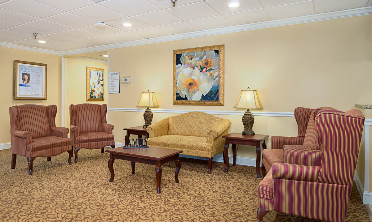 A cozy senior living facility lounge area with four striped armchairs and a yellow patterned loveseat arranged around a wooden coffee table and two side tables with decorative lamps. The walls are painted light yellow with white trim, and there are framed floral paintings and a directory sign on the walls. The carpet has a brown and beige pattern.