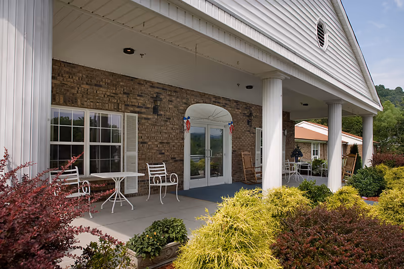 Front porch area of a building with white columns, brick walls, and glass double doors decorated with red, white, and blue ribbons. White metal chairs and tables are arranged on the porch, surrounded by green and red shrubs and plants under a partly cloudy sky.