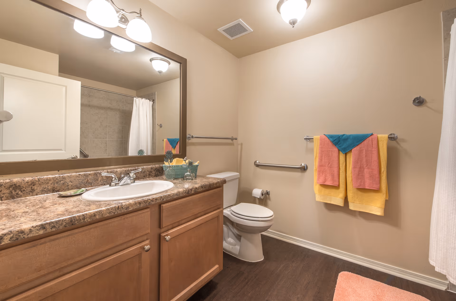 Bathroom interior with a sink and countertop, large mirror, toilet, shower curtain, and colorful towels on a rack.