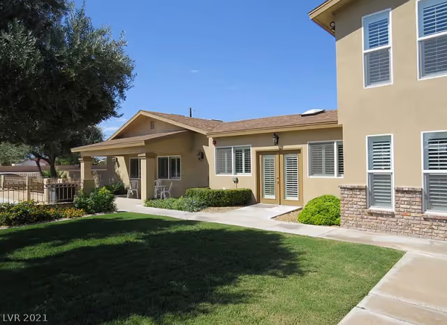 Exterior view of a beige senior living facility building with multiple windows and a small covered patio area with chairs. The building is surrounded by a well-maintained lawn, shrubs, and a large tree under a clear blue sky.