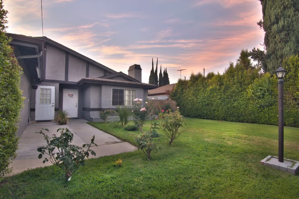 A well-maintained backyard garden area of a residential building at sunset, featuring a green lawn, several rose bushes, a paved walkway, tall hedges along the perimeter, and a lamp post on the right side.