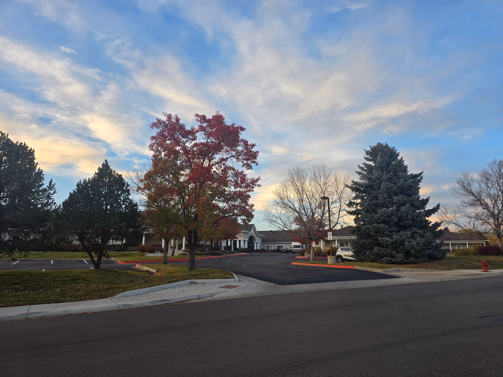 Entrance and front exterior of a senior living facility with a driveway, parked cars, and trees under a colorful sky.