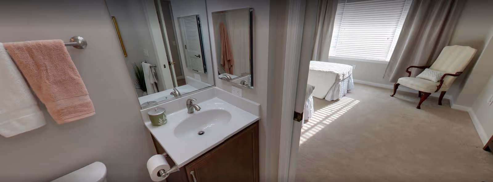 View from a bathroom into a sunlit bedroom showing a sink vanity, towels, a toilet, a bed, and an armchair.