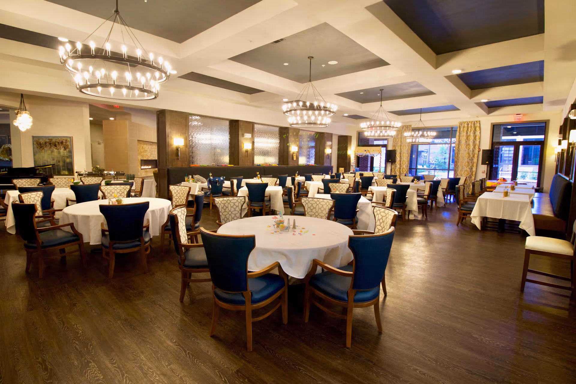 A spacious dining room with multiple round tables covered with white tablecloths and surrounded by wooden chairs with blue and patterned upholstery. The room features modern chandeliers hanging from a coffered ceiling, large windows with curtains, and a polished wooden floor. The ambiance is warm and inviting, suitable for communal dining.