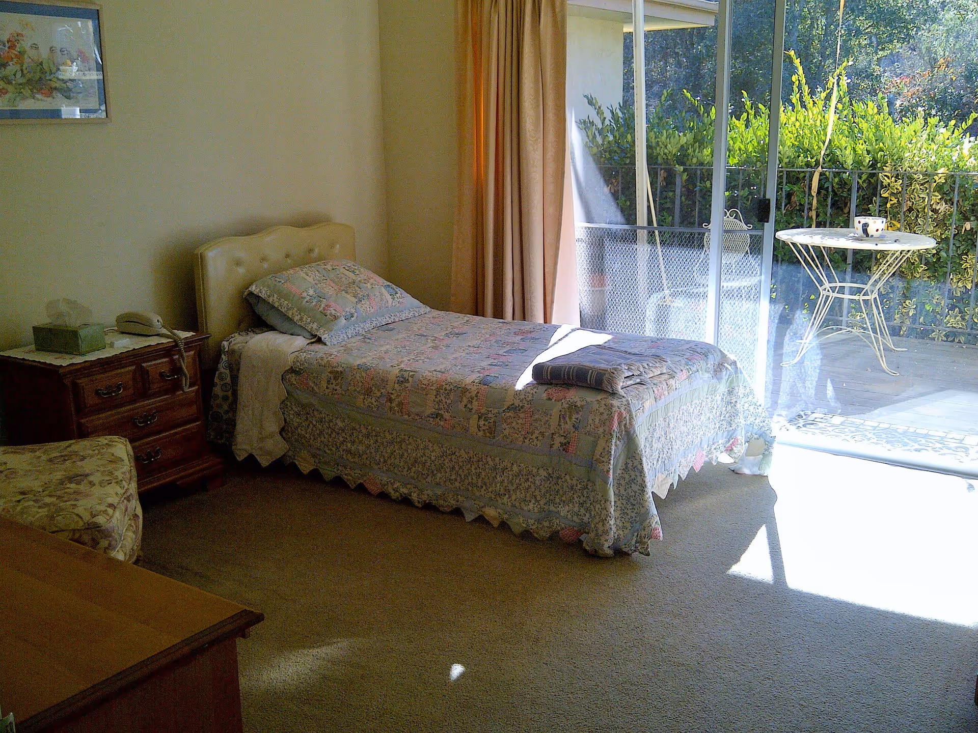 A cozy bedroom with a single bed covered in a floral quilt and pillow. Next to the bed is a wooden nightstand with a telephone and a tissue box. The room has beige walls and carpet, with a sliding glass door leading to an outdoor patio area with a small round table and chair surrounded by greenery.
