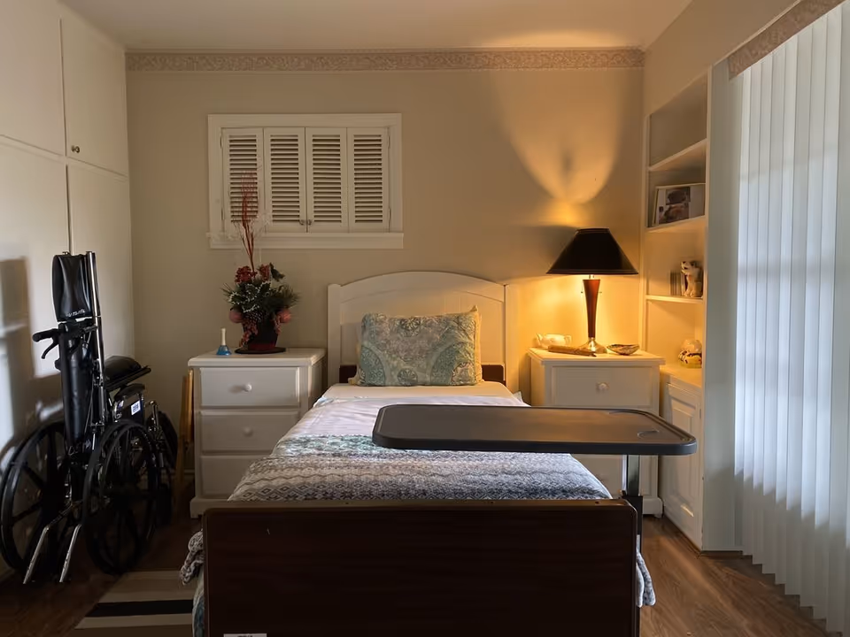 A small, cozy bedroom with a single bed centered between two white nightstands. The bed has a patterned quilt and a decorative pillow. A black wheelchair is positioned to the left side of the room. On the right nightstand, there is a lamp with a dark shade casting warm light. Built-in shelves with decorative items and a window with vertical blinds are on the right wall. The walls are beige, and the floor is wooden.