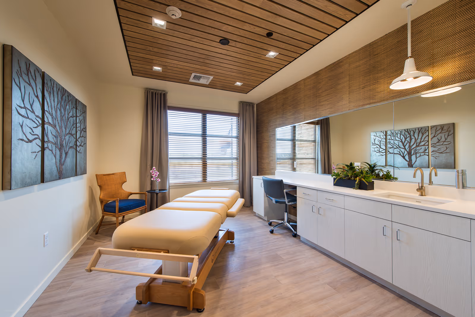 A well-lit therapy or treatment room with a padded treatment table in the center, a wooden chair with a blue cushion next to a small round table holding a pink orchid, a large mirror above a long white cabinet with a sink and faucet, and a wooden ceiling with recessed lighting. A three-panel artwork of a tree is mounted on the wall.