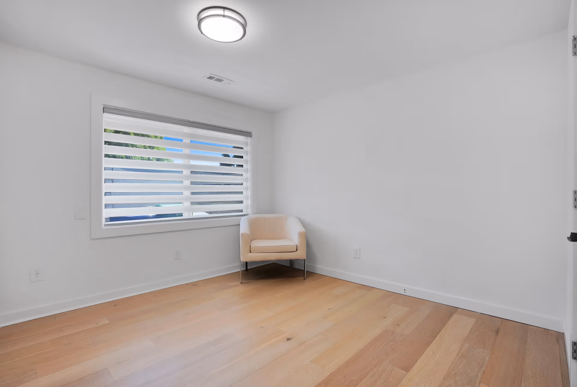 A small, empty room with light wooden flooring and white walls. There is a single beige armchair placed in the corner near a window with horizontal blinds. A round ceiling light fixture is mounted on the white ceiling.