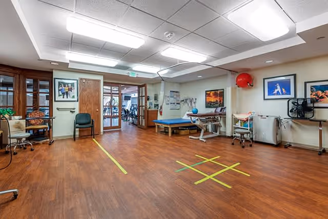 A spacious room with wooden flooring and ceiling lights, featuring exercise and therapy equipment including a treatment table, exercise ball, and a tic-tac-toe game marked on the floor with tape. There are chairs, framed pictures on the walls, and a doorway leading to another room with more exercise equipment visible.