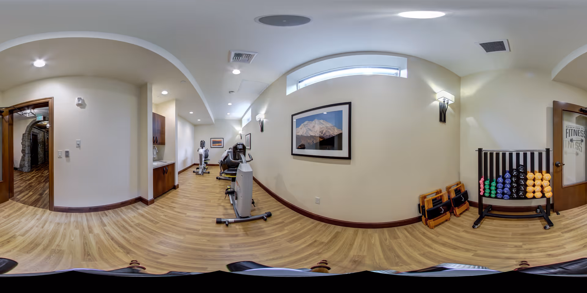 A fitness room with exercise equipment including stationary bikes and a rack of colorful dumbbells. The room has wooden flooring, cream-colored walls, wall-mounted lights, and a framed picture of a mountain. There is a door labeled 'Fitness' and a small sink area with cabinets.