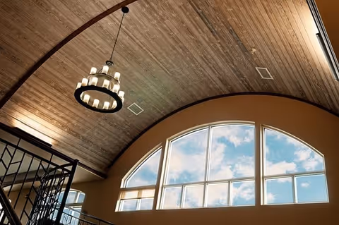 Interior view of a room with a high, curved wooden ceiling featuring a circular chandelier with multiple lights. Below the ceiling is a large arched window showing a blue sky with clouds. Part of a black metal railing is visible on the left side.